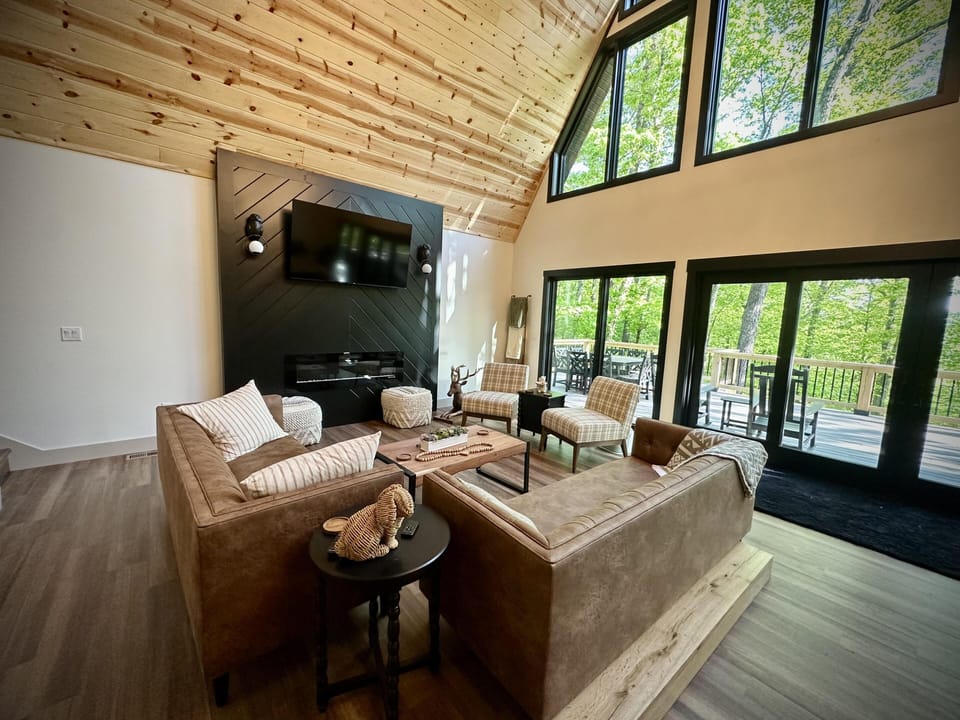 Living room features two tan leather sofas, plaid armchairs, black paneled media wall, and floor-to-ceiling windows.