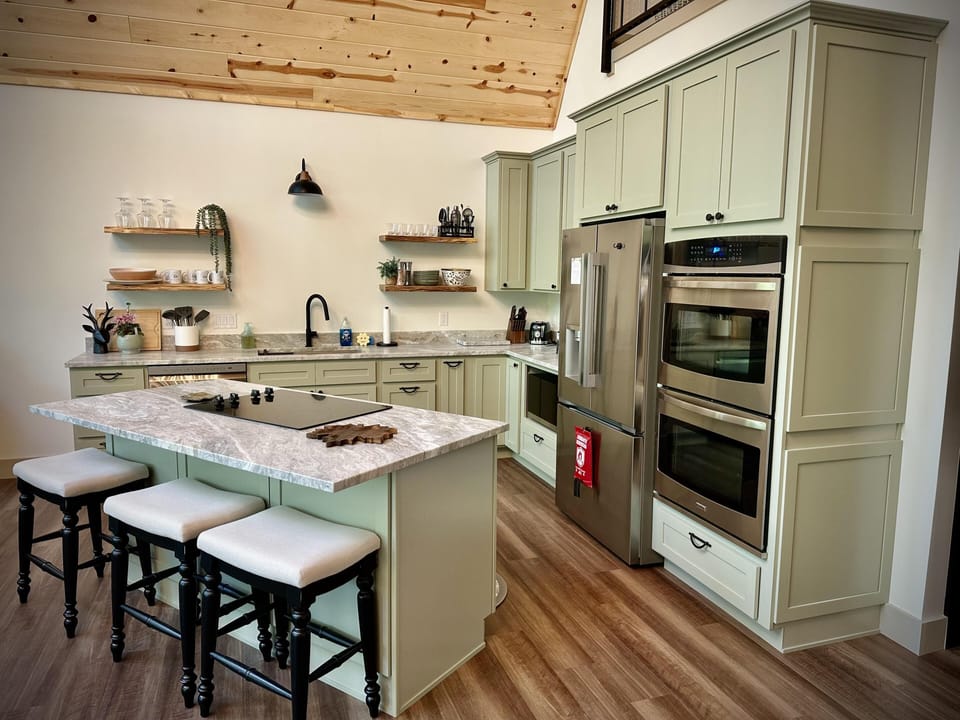 Kitchen with sage green cabinetry, marble countertops, stainless steel double ovens, and black faucet under vaulted wood ceiling.