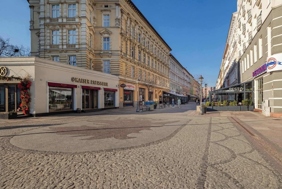 A city street with elegant buildings and shops along the sides. The perspective highlights the depth and charm of the neighborhood.