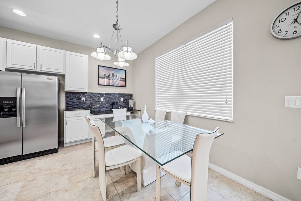 This bright kitchen features sleek white cabinetry with dark accents, a stylish glass dining table for gatherings, and plenty of natural light from a large window, creating a warm and inviting space for you and your guests.