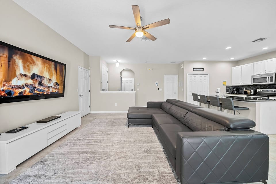 Relax in the spacious living room, where a sleek black sectional sofa invites comfort. The modern wall fireplace adds warmth, and the ceiling fan along with natural light creates a bright, airy atmosphere for your enjoyment.