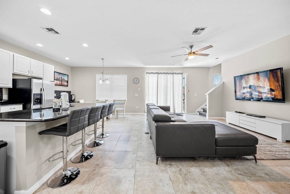 Experience a bright living area with a sleek leather sectional and a large flat-screen TV, ideal for relaxing evenings. The open layout seamlessly connects to a stylish kitchen with modern bar stools, perfect for gatherings.