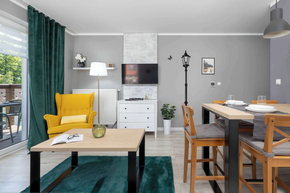 A view of the apartment’s interior connecting the dining room with the living room, where a contrasting yellow armchair stands next to the wooden table. The light-colored walls harmonize with the elegant hanging curtains and modern lighting.