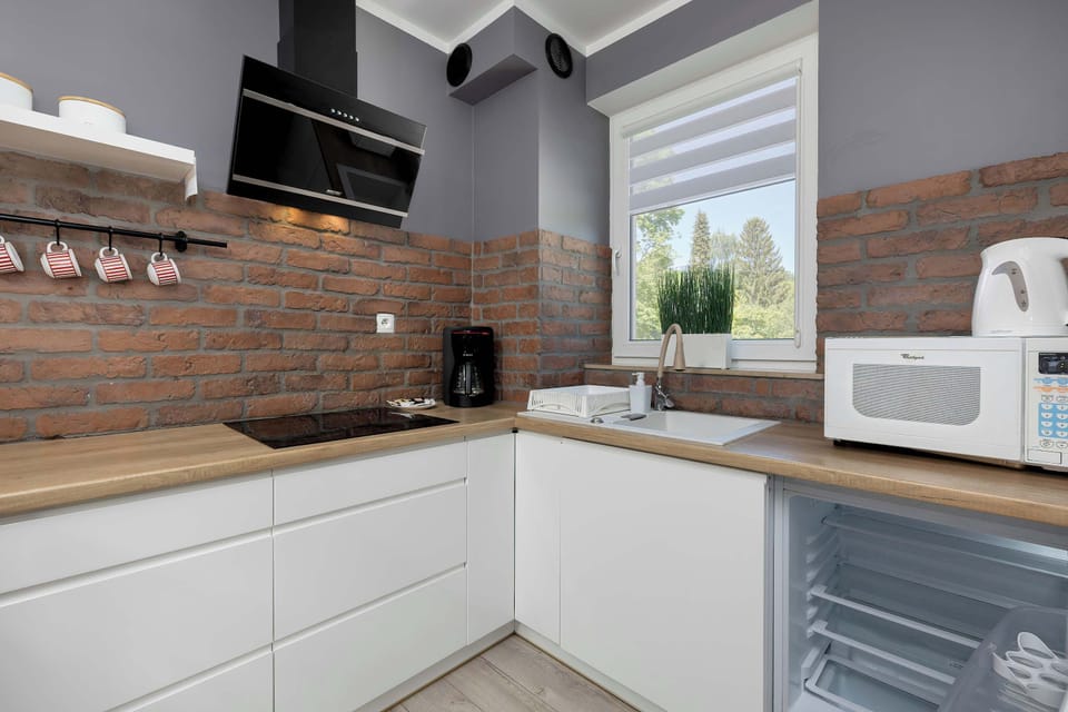 A close-up of the corner kitchen cabinets with white fronts and a natural wood-colored countertop, contrasting with the dark brick wall. The countertop holds essential appliances, including a kettle, coffee maker, and microwave.