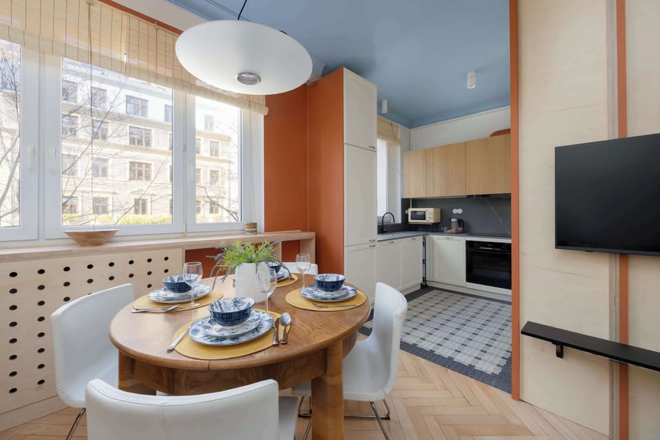 A bright, open-plan living area features a round wooden dining table set with blue patterned bowls and four white chairs positioned near a large window. A vibrant orange accent wall separates the dining space from a modern kitchen equipped with wood 