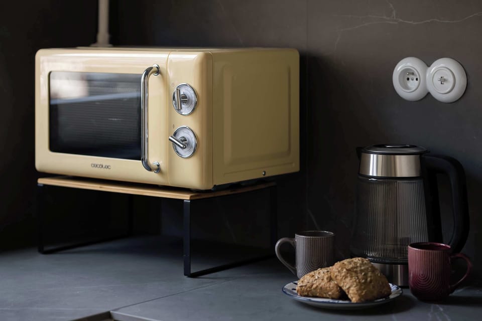 A detailed shot of a retro-style yellow toaster oven on a black stand, with a sleek black kettle and a plate of baked goods nearby on a dark surface, against a dark wall with a decorative circular detail.