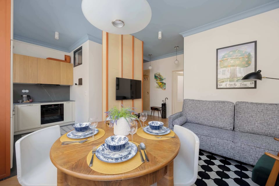A smaller-framed picture of a dining area featuring a round table with blue-rimmed plates and white chairs, positioned near a window, with a partial view of a sofa and blue ceiling trim.