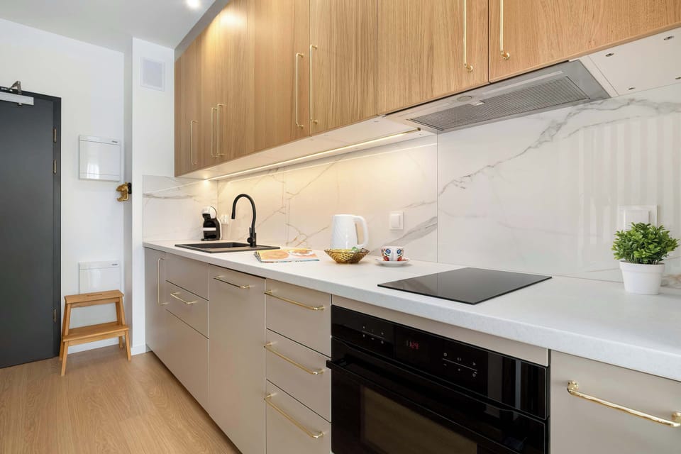 This L-shaped kitchen maximizes efficiency with a built-in oven, induction cooktop, and ample counter space. Light wood tones and marble-patterned backsplashes provide a modern, airy feel.