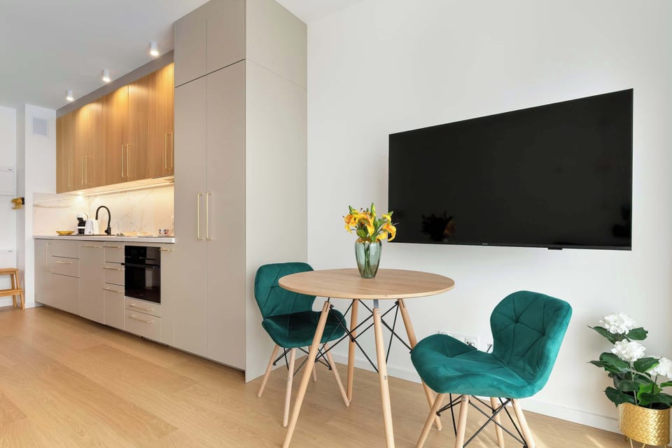 This open-concept living area seamlessly connects a compact dining set with two teal chairs to a modern kitchen. A flat-screen TV is mounted on the wall, making the space perfect for both dining and entertainment.