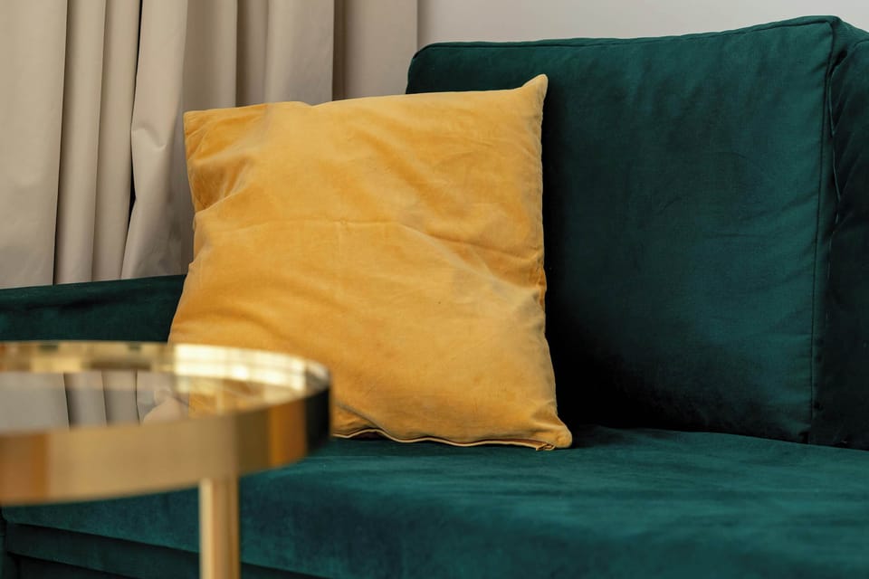 A plush yellow pillow provides a bright pop of color against the deep teal velvet of a contemporary sofa. In the foreground, the edge of a polished gold coffee table catches the light.