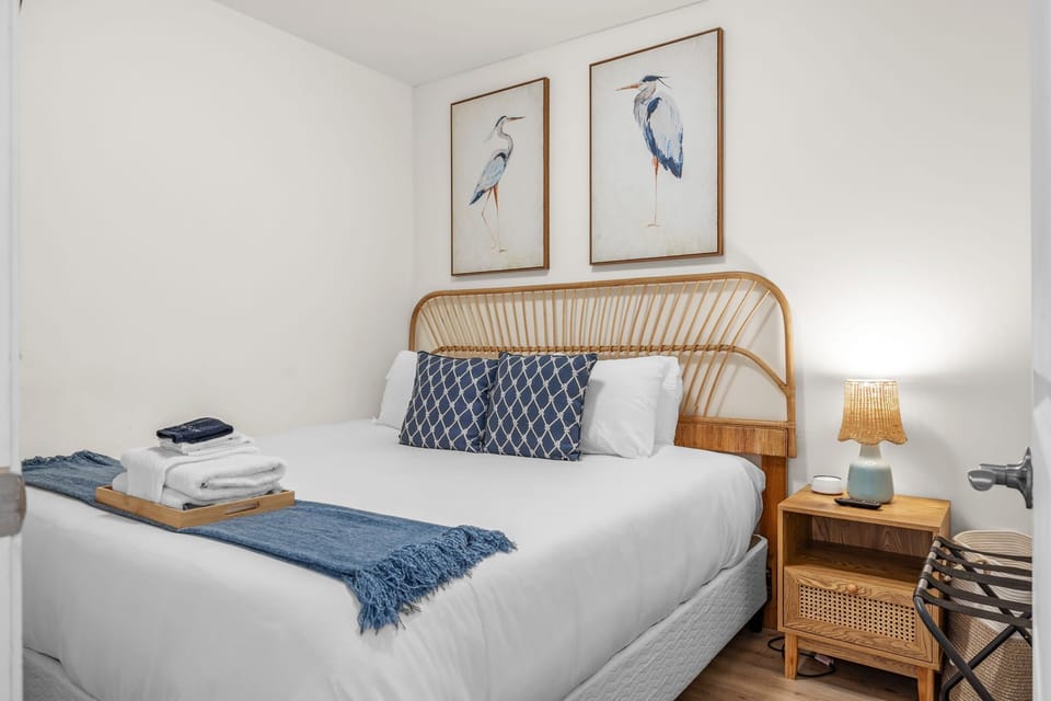 Bedroom with a woven headboard, heron artwork, white linens, blue accents, and bedside lighting.