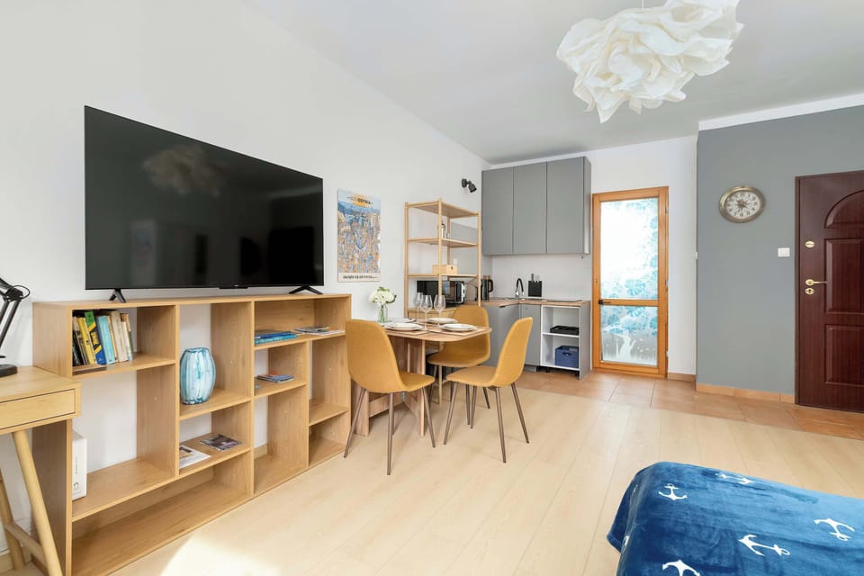 A wider view of the studio showing the bed, desk with chairs, and dining table in one open area. The layout is neatly organized with modern furniture and wooden floors.