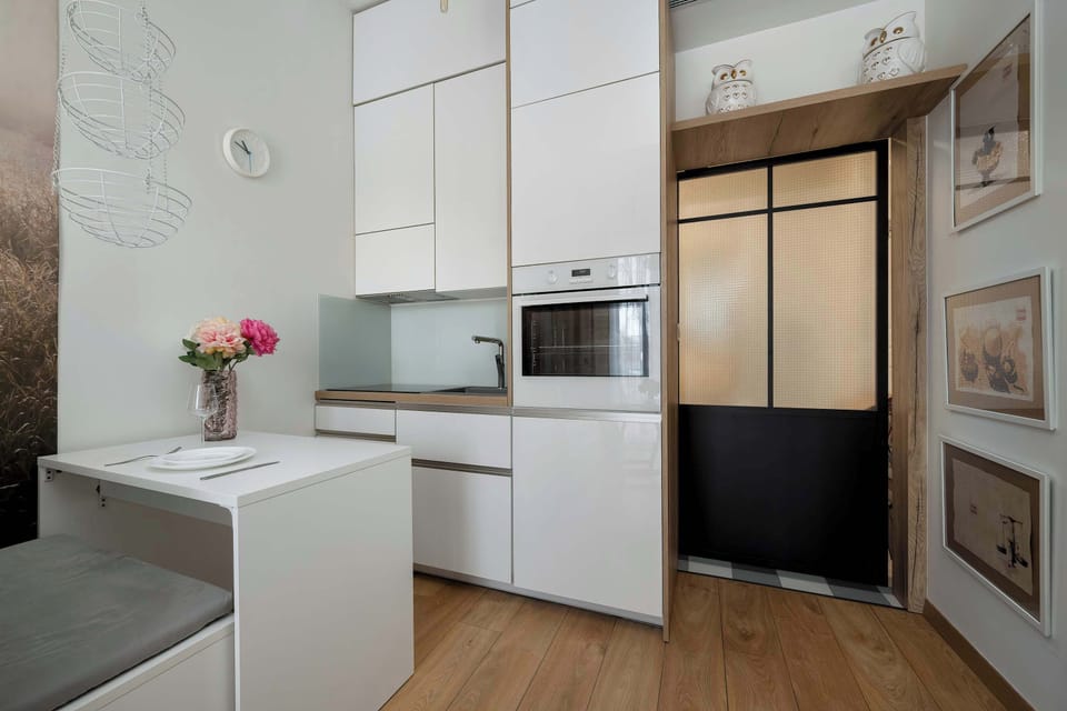 A functional dining area includes a small white table set with a pink floral centerpiece. The compact kitchenette is visible in the background, featuring integrated appliances and sleek white finishes.