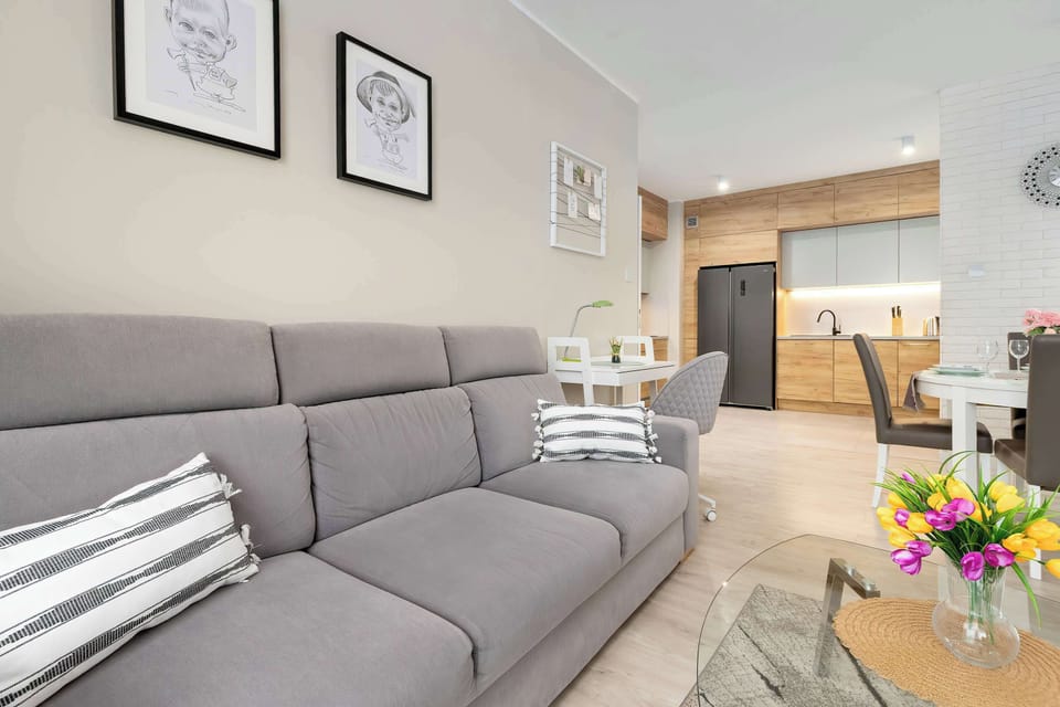 The camera looks across the grey sofa toward the kitchen, showing the seamless flow between the living and cooking areas. Bright floral arrangements are placed on both the coffee and dining tables.