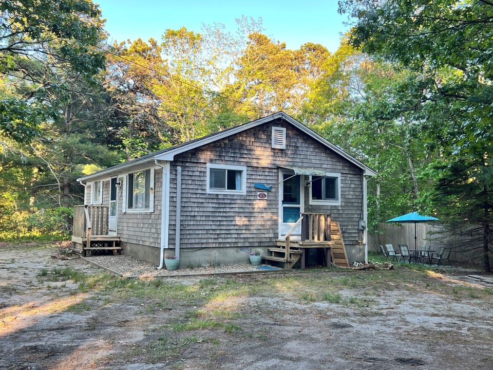 Charming shingled cottage nestled among mature trees with outdoor seating area for peaceful woodland relaxation.