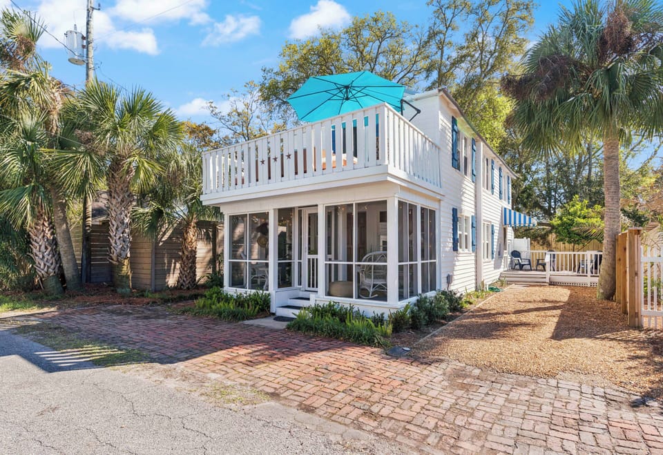Charming coastal cottage featuring a wraparound porch and upper deck with turquoise umbrella, surrounded by palm trees and tropical landscaping.