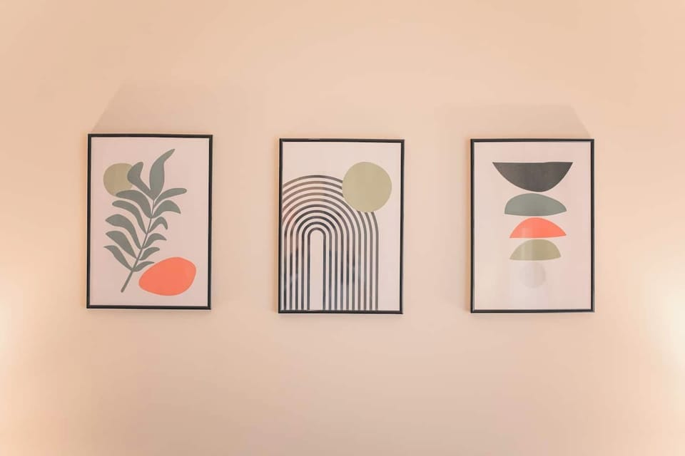 A minimalist trio of framed abstract art pieces decorates the wall. Each print features earthy tones of green, orange, and black with simple shapes—leaves, arches, and stacked forms—adding a modern and calming aesthetic to the space.