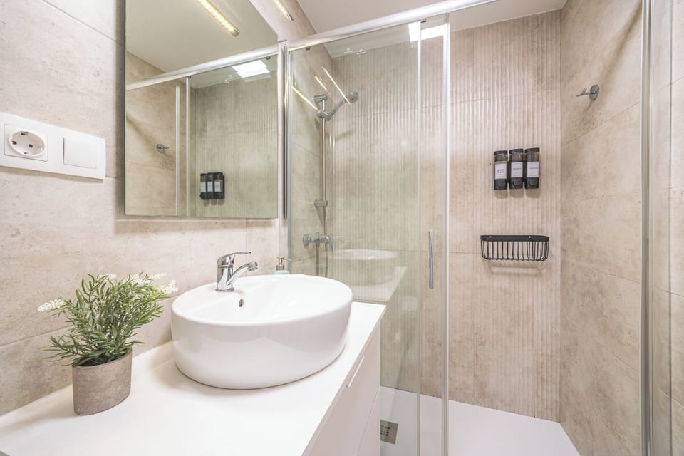 A bright bathroom with a sink with cabinets, a hanging mirror, and a large shower with dispensers, a coat rack, and a shelf.