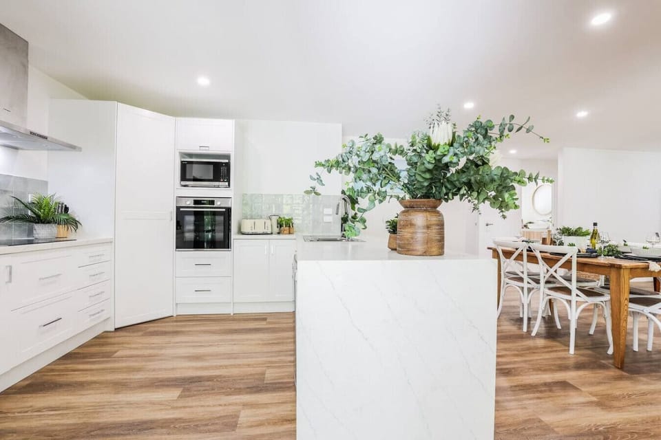 Open‑plan kitchen with marble island, stainless appliances, timber dining table, and coastal greenery accents. Family‑friendly dining space.