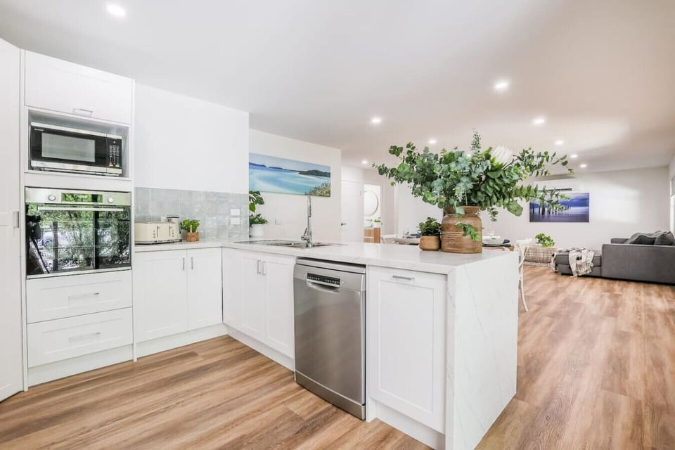 Modern kitchen with white cabinetry, stainless appliances, marble‑finish counters, and timber flooring. Open‑plan living with gray sofa and coastal artwork.