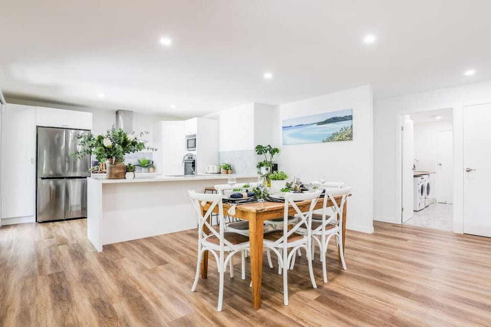 Modern kitchen with white cabinetry, stainless appliances, timber dining table, and woven chairs. Family‑friendly dining space.