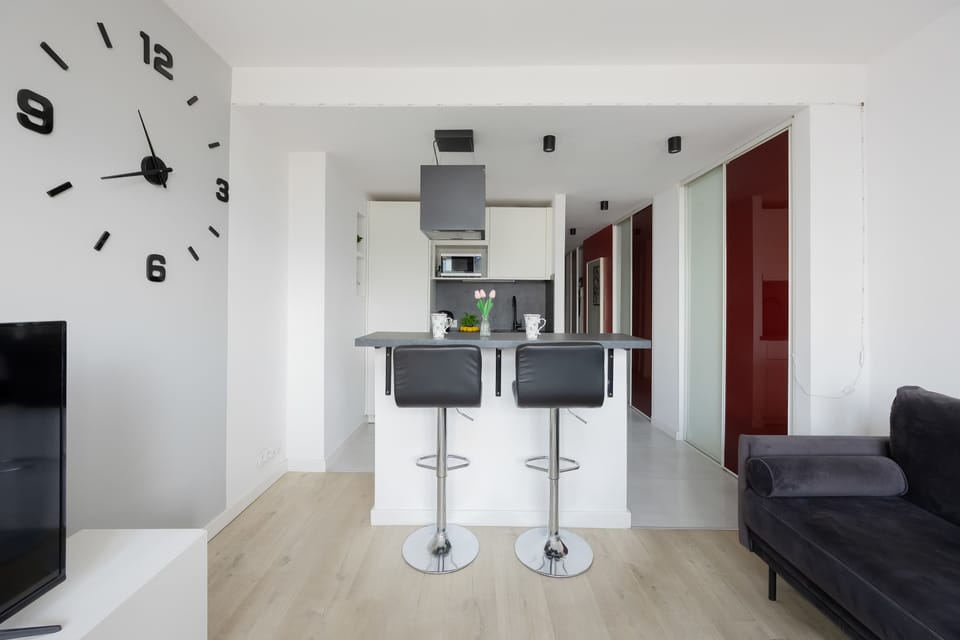 kitchen area with a breakfast bar and two white bar stools. The space is clean and contemporary, with white cabinets and subtle decorative elements. A wall clock adds a stylish touch.