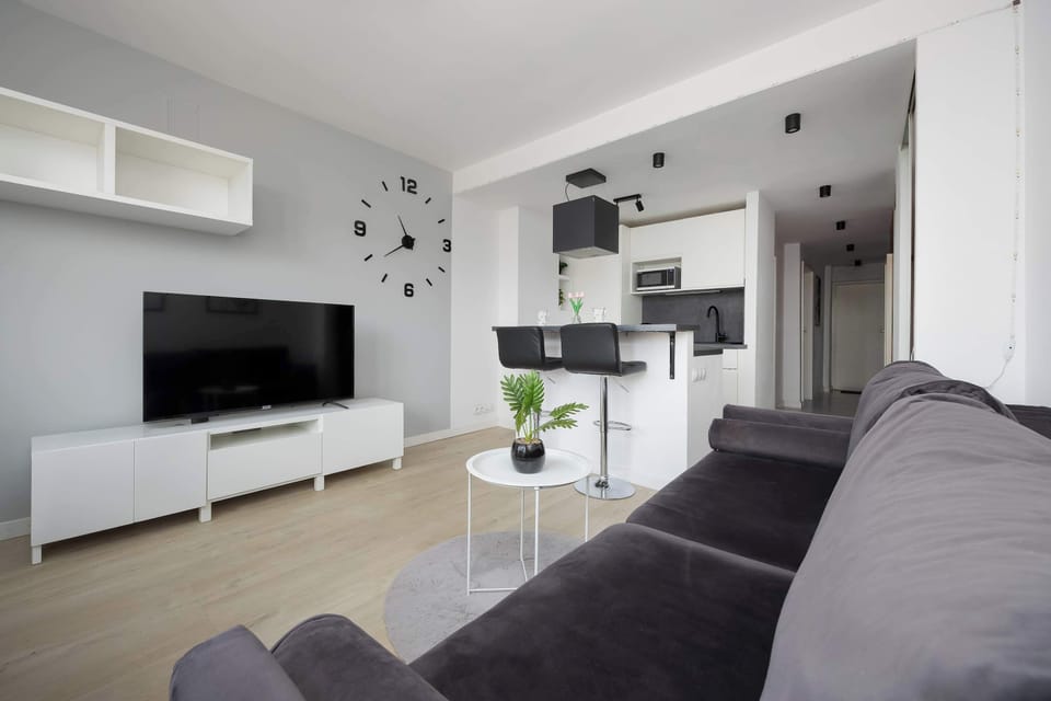 A modern open-plan living room connected to a kitchenette. A gray corner sofa faces a TV, while the kitchen area is sleek and fully equipped. Neutral tones dominate the interior.