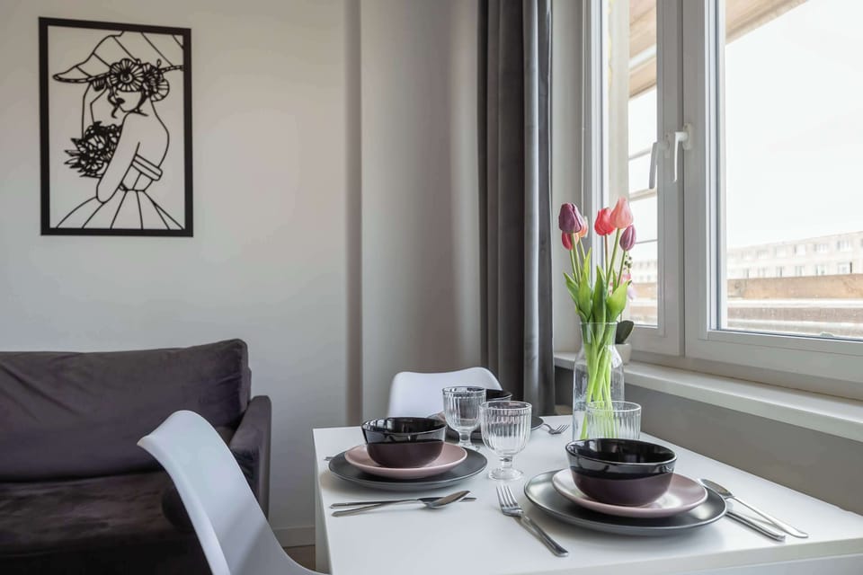 A close-up of a dining setup by the window with plates, cups, and decorative flowers. Natural light highlights the table setting. The arrangement feels inviting and homely.