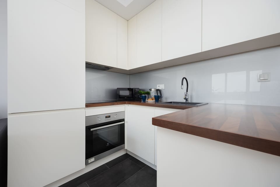 A modern kitchenette setup showcases white handleless cabinetry and a dark wood-effect countertop. The space is equipped with an integrated oven and a minimalist black sink.