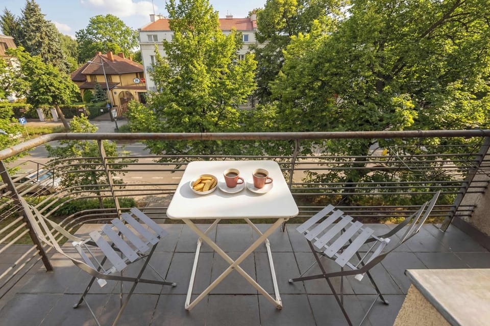 A spacious balcony is furnished with a wooden table and chairs, perfect for outdoor relaxation. It offers an elevated view of the surrounding urban landscape under a clear sky.