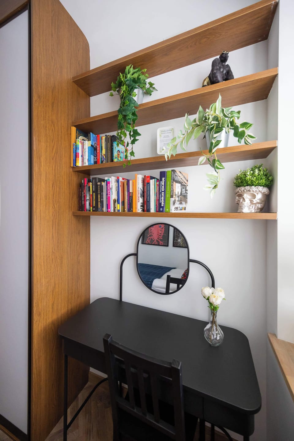 A compact vanity area includes a small desk and a circular mirror, flanked by wooden shelving units. Potted plants and books are scattered throughout, adding a lived-in feel to the nook.