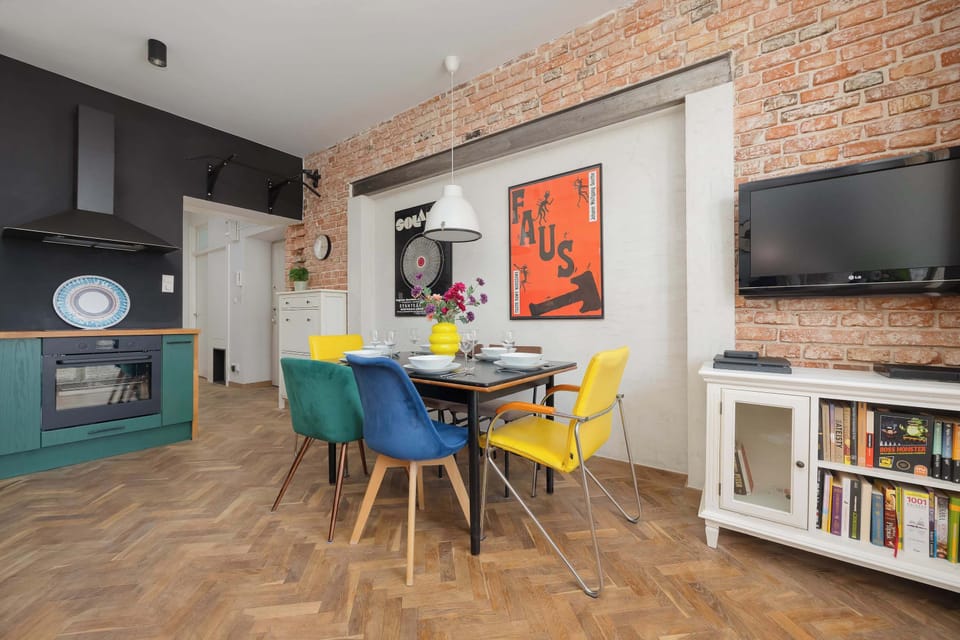 A bright, open dining space contains a large table and a collection of mismatched colorful chairs. An exposed brick wall with stylized posters forms a striking backdrop for family meals.