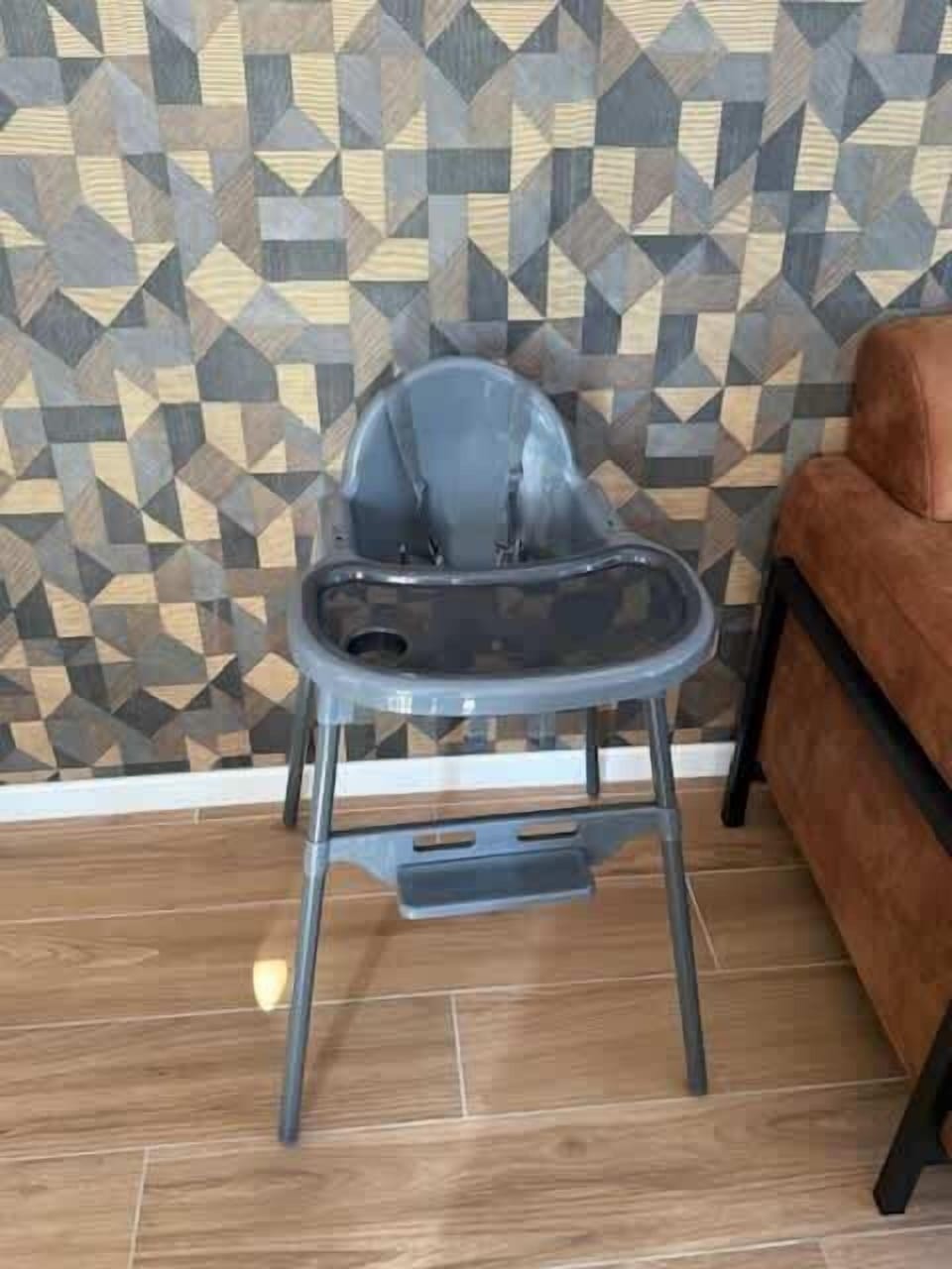 A functional gray high chair placed next to a brown sofa against a geometric patterned wall.