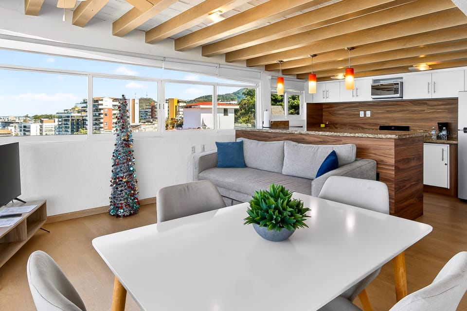 Modern white dining table with grey chairs, seamlessly connected to the living room and bordered by bright windows.