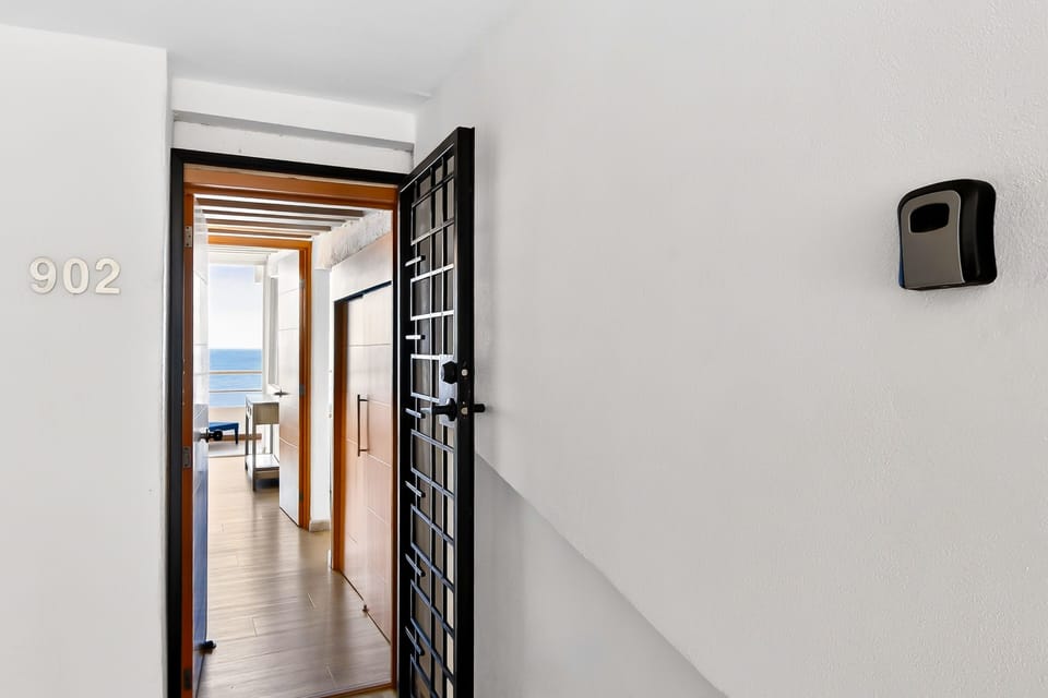 View of the open black metal security gate and front door, revealing a bright hallway path that leads straight toward the ocean.