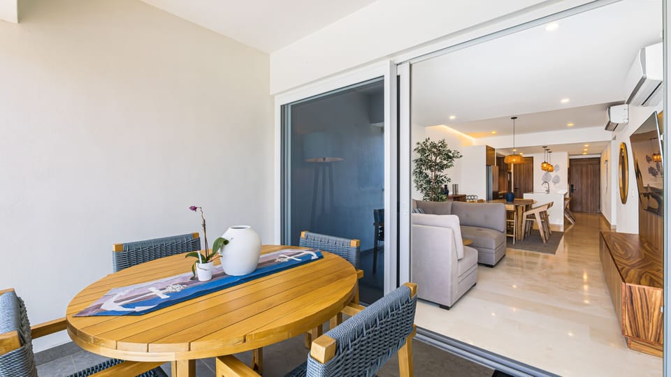 Oval wooden dining table with a nautical table runner set on a private terrace, looking through open sliding glass doors into the main open-concept apartment space.