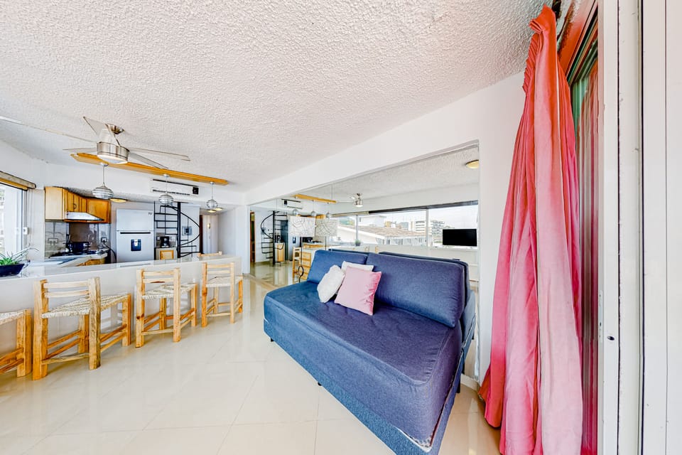 Comfortable living space highlighting a deep blue sofa bed with pink accent pillows, opening up to the kitchen bar area.