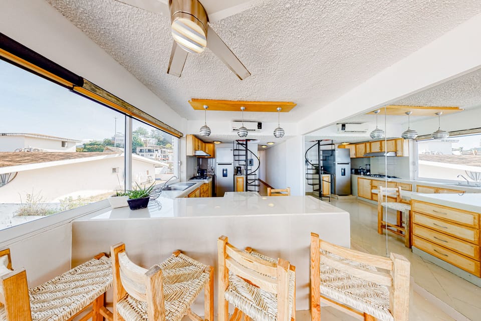 Neat dining counter setup highlighting four woven wooden bar stools, modern hanging pendant lights, and bright surrounding windows.