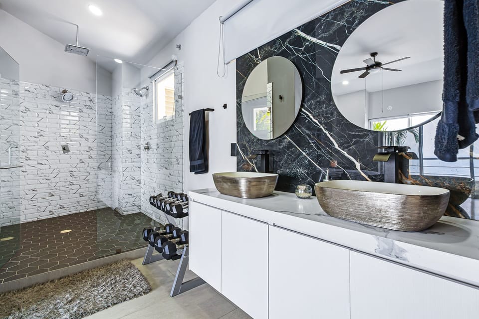 A bright en-suite bathroom view highlighting dual vessel sinks, dark marble accents, and a spacious glass-enclosed walk-in shower with marble-look subway tiles.