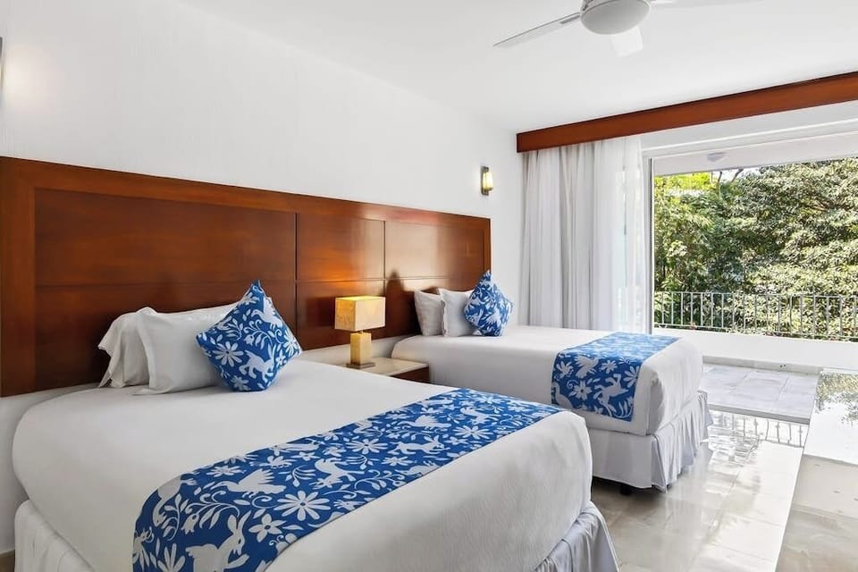 A well-lit bedroom featuring two beds with white linens, blue decorative runners, a wooden headboard, and balcony views.