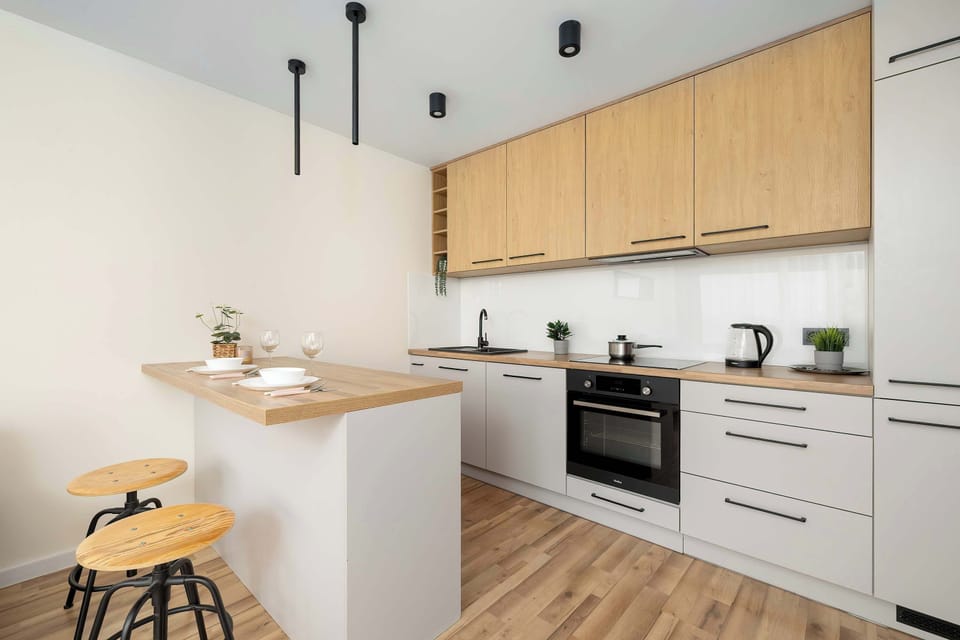 A modern kitchenette with light-colored cabinet fronts and a wooden countertop, equipped with an induction cooktop, an oven, and a bar stool at the breakfast island.