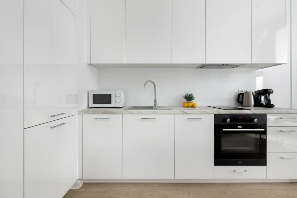 A close-up view of a sleek, white-cabinetry kitchen with a light-colored countertop. It features an integrated black oven, a microwave, a small sink, and an induction cooktop.