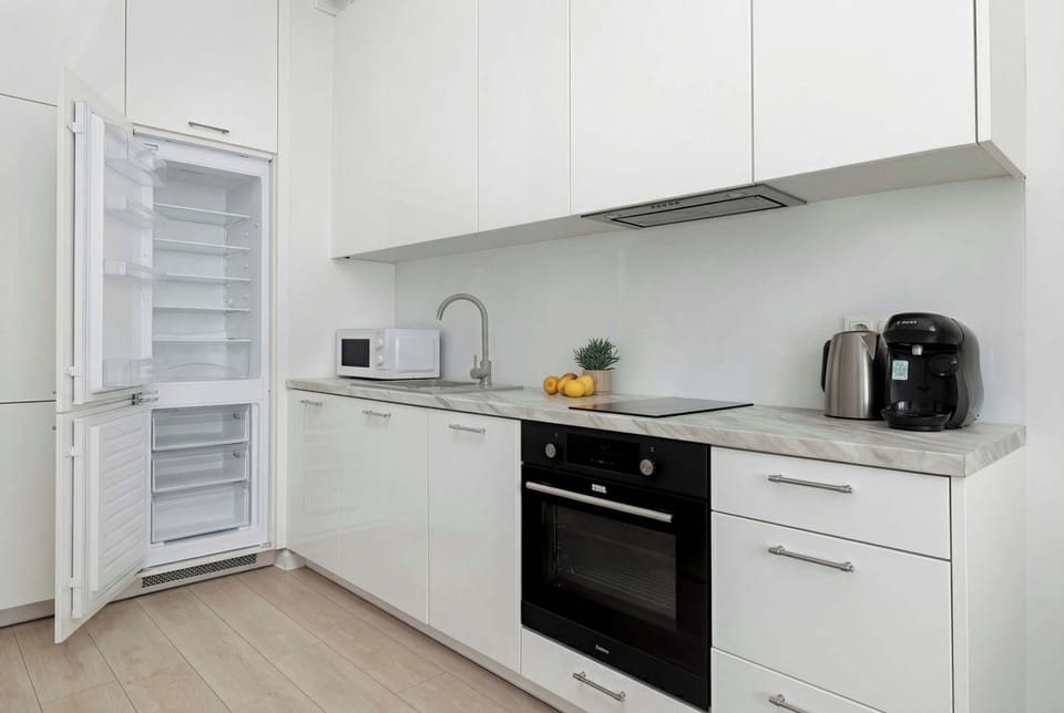 A kitchen scene showcasing the open refrigerator to the far left. The rest of the kitchen features seamless white cabinetry, an integrated black oven, and a dark sink.