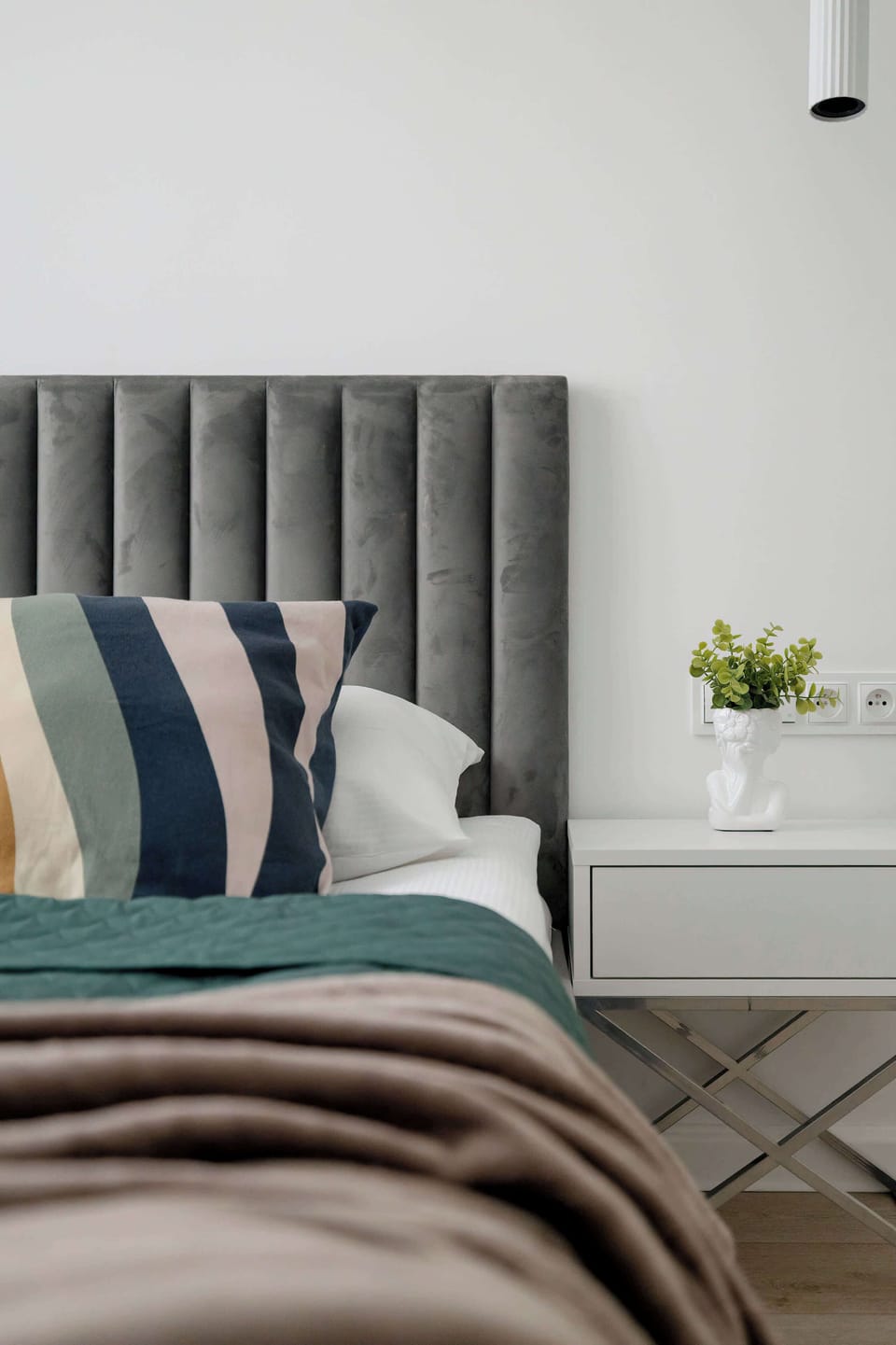A vertical close-up shot focused tightly on a bed, showing the grey tufted headboard. A patterned pillow with blue stripes sits on top of a dark teal duvet.