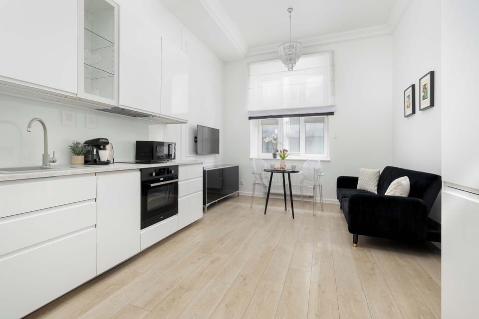 A wide perspective of the apartment shows the open-plan layout connecting the kitchen, dining area, and lounge. Large windows at the far end fill the entire space with natural light, highlighting the uniform wooden floors.