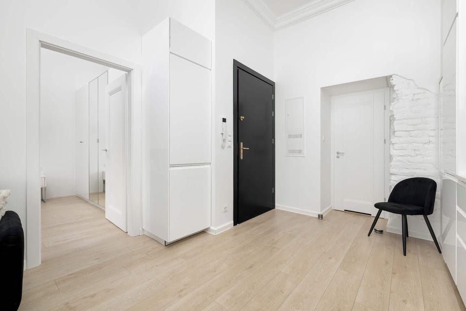A spacious hallway area features several white doors and a large built-in wardrobe for guest storage. A black entrance door and a simple black chair maintain the apartment's consistent color palette.
