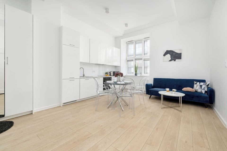 A view of the living room with a kitchenette, showing a large window that lets in plenty of natural light. The entire space is decorated in a cohesive white-and-blue color scheme with modern accents.