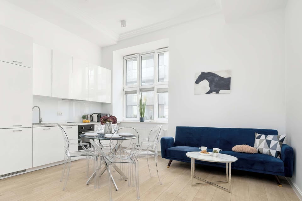 A spacious kitchenette connected to the living room, dominated by modern, white cabinetry. The seating area features a deep, blue sofa, next to which stands a glass table with designer chairs.