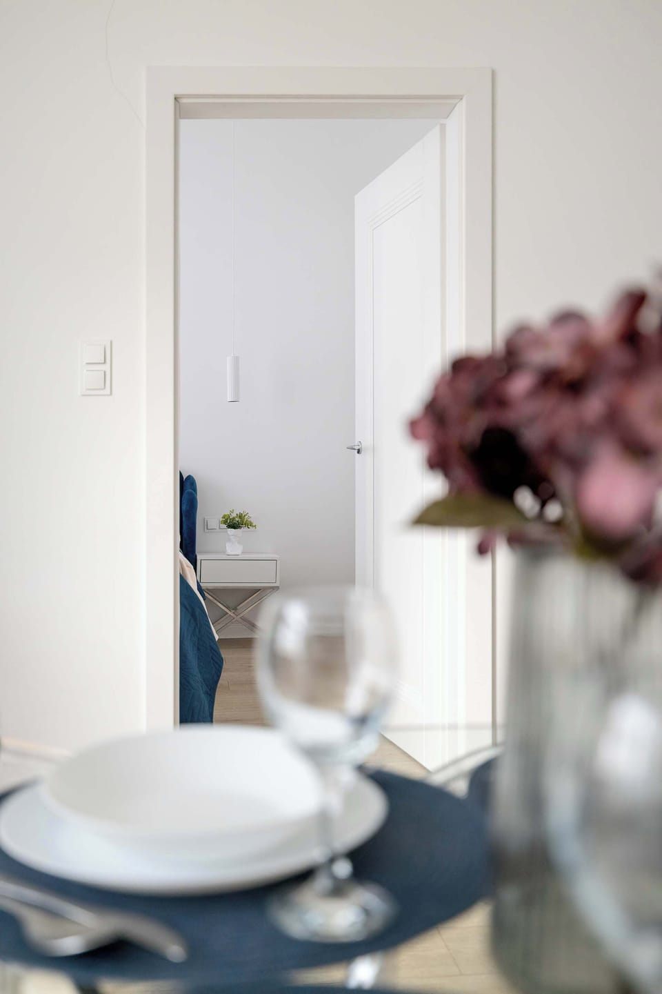 A decorative detail featuring a white plate on a dark coaster, placed on a glass dining table. Fresh flowers are visible in the background, adding a touch of homey warmth and elegance to the interior.