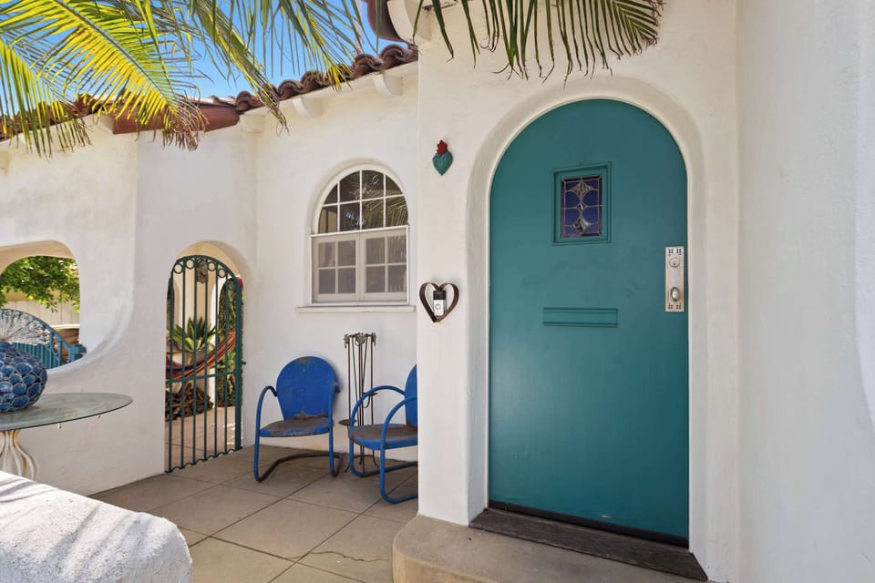The covered front entry porch — teal arched door with stained glass inset, vintage blue metal chairs, and an ornamental iron gate opening to the garden. A welcoming arrival in true LA style.